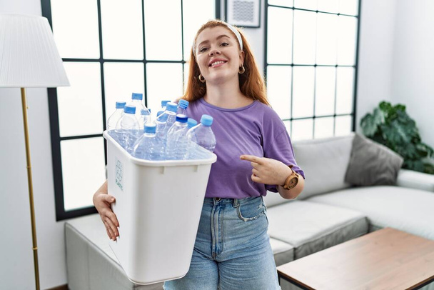 Junge rothaarige Frau mit Recycling-Papierkorb und Plastikflaschen, die mit dem Zeigefinger zur Seite zeigen und Werbung zeigen, ernstes und ruhiges Gesicht  - Foto, Bild