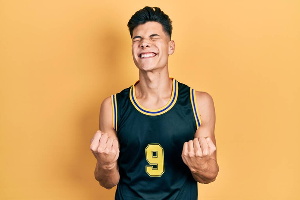 Junger hispanischer Mann in Basketballuniform sehr glücklich und aufgeregt dabei Siegergeste mit erhobenen Armen, lächelnd und schreiend nach Erfolg. Festkonzept.  - Foto, Bild