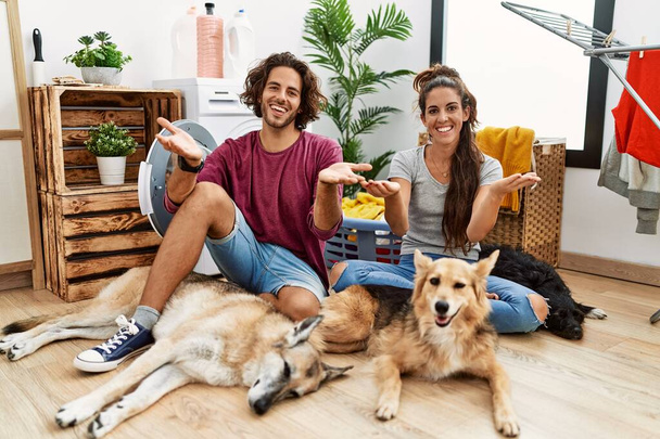 Young hispanic couple doing laundry with dogs smiling cheerful offering hands giving assistance and acceptance.  - Photo, image