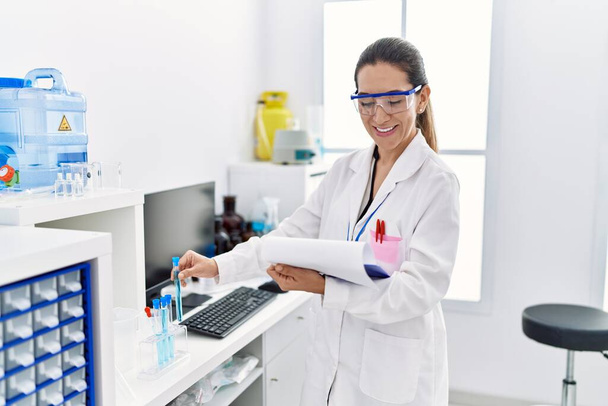 Laboratuvarda deney tüpü tutan bilim adamı üniforması giyen İspanyol genç bir kadın. - Fotoğraf, Görsel