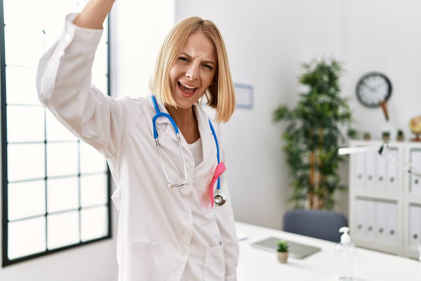 Jovem mulher médica caucasiana vestindo fita de câncer rosa irritado e frustrado gritando com raiva, gritando louco com raiva e mão levantada  - Foto, Imagem