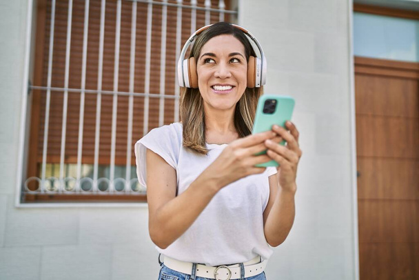 Mladá hispánka se usmívá sebevědomě pomocí smartphonu a sluchátek na ulici - Fotografie, Obrázek