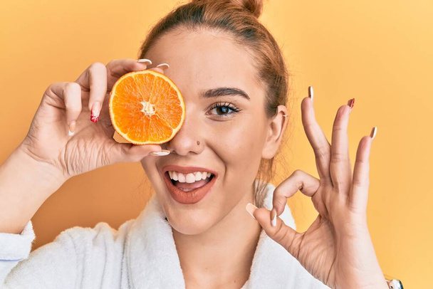 Jeune femme blonde portant un peignoir tenant orange frais faisant signe ok avec les doigts, souriant geste amical excellent symbole  - Photo, image