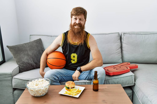 Homme caucasien à longue barbe tenant ballon de basket-ball acclamant jeu de télévision clin d'oeil en regardant la caméra avec une expression sexy, gai et heureux visage.  - Photo, image