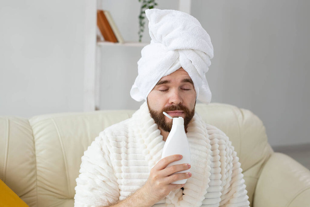 Bearded male with towel on his head applying body lotion cream. Guy smile when smell lotion cream on hand. Spa, body and skin care for man concept - Photo, Image