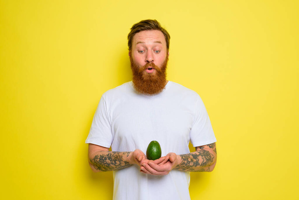 Erstaunt mit Bart und Tattoos hält eine Avocado in der Hand - Foto, Bild