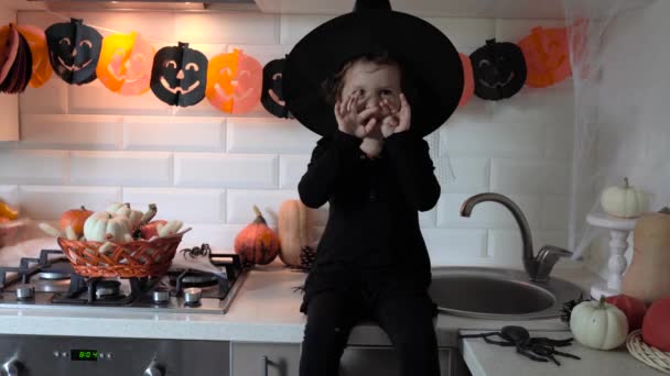 Niña en traje de Halloween bruja jugando con decoraciones y sentado en la mesa de la cocina con calabazas, guirnalda y arañas - Metraje, vídeo