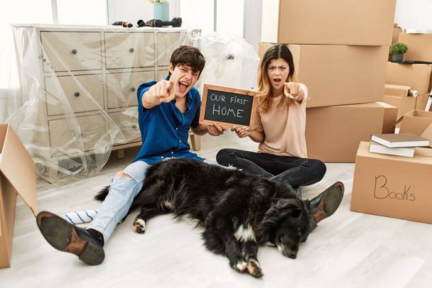 Young caucasian couple with dog holding our first home blackboard at new house pointing displeased and frustrated to the camera, angry and furious with you  - Foto, Imagem