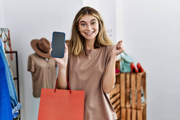 Mujer rubia joven sosteniendo bolsas de compras mostrando la pantalla del teléfono inteligente gritando orgulloso, celebrando la victoria y el éxito muy emocionado con el brazo levantado  - Foto, Imagen