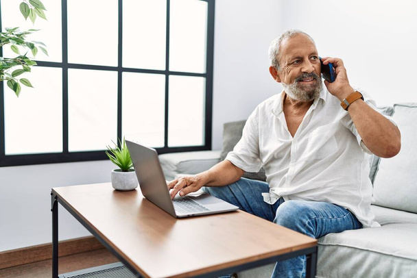 Senior hombre de pelo gris hablando en el teléfono inteligente usando el ordenador portátil en casa - Foto, imagen