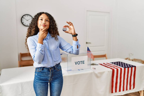 Beautiful hispanic woman standing by at political campaign by voting ballot looking confident at the camera smiling with crossed arms and hand raised on chin. thinking positive.  - Photo, Image