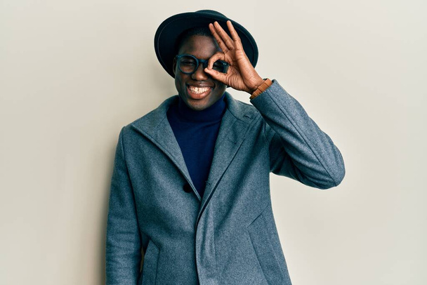 Junger afrikanisch-amerikanischer Mann trägt eleganten Stil und macht ok Geste mit lächelnden Händen, Auge blickt durch Finger mit glücklichem Gesicht.  - Foto, Bild