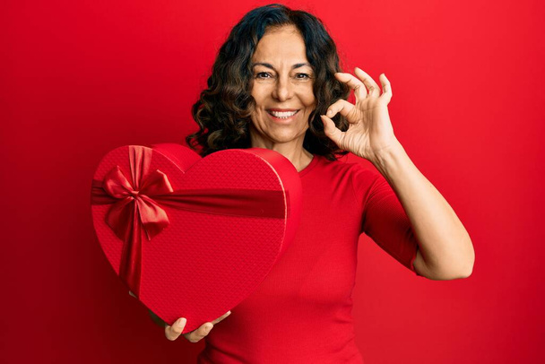 Mujer hispana de mediana edad sosteniendo regalo de San Valentín haciendo signo de ok con los dedos, sonriendo gesto amistoso excelente símbolo  - Foto, imagen