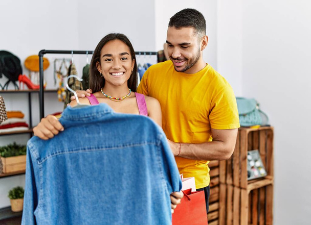 Młoda Latynoska para uśmiechnięta szczęśliwa wybierając ubrania w sklepie odzieżowym. - Zdjęcie, obraz