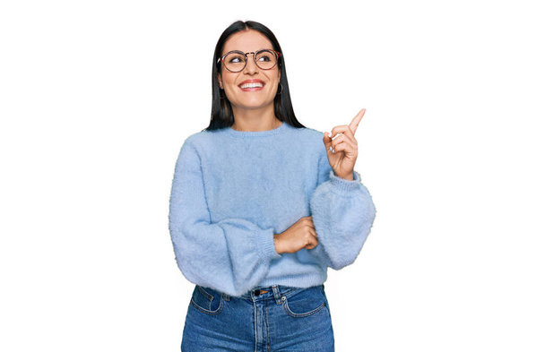 Молодая латиноамериканка в повседневной одежде и очках с широкой улыбкой на лице, указывая рукой и пальцем в сторону, глядя в камеру.  - Фото, изображение