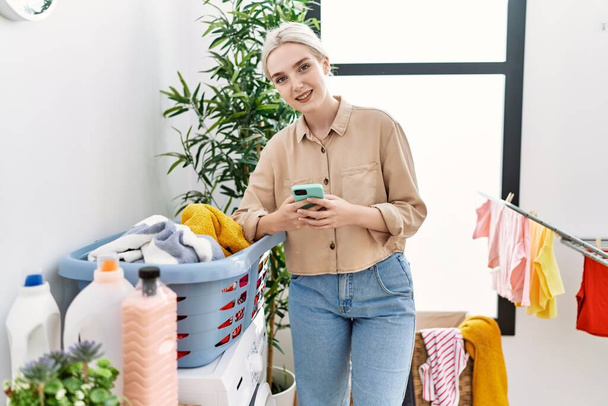 Young caucasian woman smiling confident using smartphone at laundry room - Фото, зображення