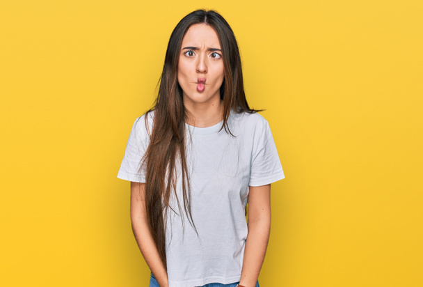 Jong Latijns-Amerikaans meisje draagt casual wit t-shirt met vissengezicht met lippen, gek en komisch gebaar. grappige uitdrukking.  - Foto, afbeelding