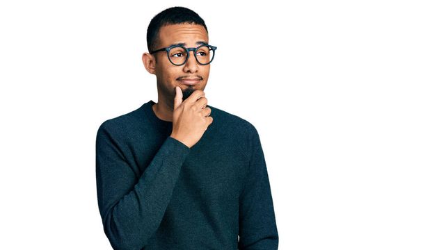 Genç Afro-Amerikan bir adam günlük kıyafetler giyiyor ve elleri çenesinde gözlük takıyor soru sormayı düşünüyor, düşünceli bir ifade. Düşünceli bir yüzle gülümsüyor. şüphe kavramı.  - Fotoğraf, Görsel