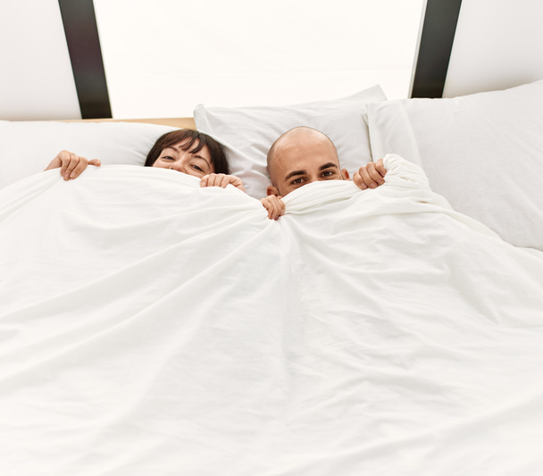 Young hispanic couple covering face with bedsheet lying on the bed at bedroom. - Foto, afbeelding