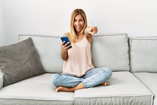 Блондинка красивая молодая женщина, сидящая дома на диване с помощью смартфона, показывающего на вас и камеру пальцами, улыбается позитивно и весело  - Фото, изображение