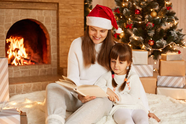 Indoor shot of mother and daughter reading fairy tails in new year eve, woman wearing white sweater and Santa Claus hat reads book her charming kid, posing in festive room on floor. - Φωτογραφία, εικόνα