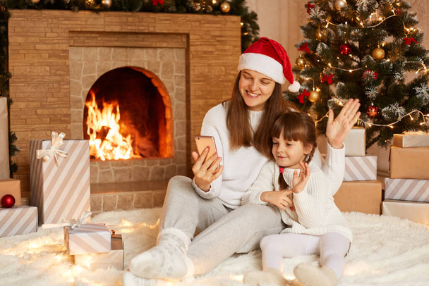 Indoor shot of mother and little daughter having video call or broadcasting livestream, waving hands to smart phone camera, posing near fireplace and Christmas tree. - Foto, immagini