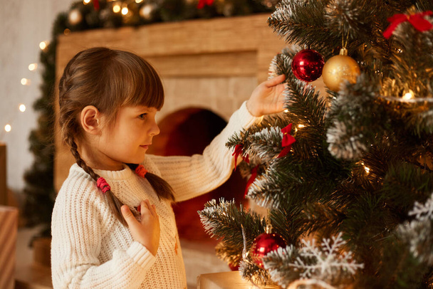 Side view portrait of charming little girl with pigtails decorating Christmas tree alone, wearing white sweater, standing in living room near fireplace. - Photo, Image