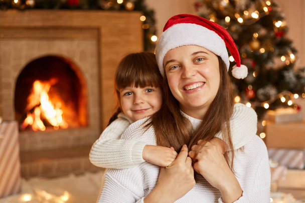 Happy family spending time together, mother and her little daughter hugging while sitting on floor in festive living room with fireplace and Christmas tree. - Fotoğraf, Görsel
