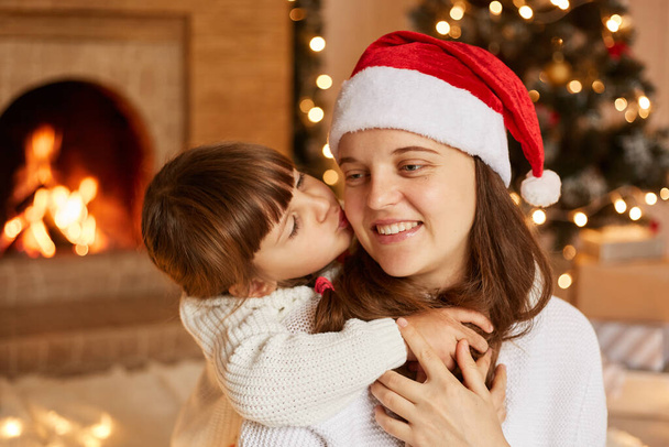 Indoor shot of mother and her little daughter hugging each other, having good mood, little cute girl kissing her mommy, merry Christmas happy new year. - Fotografie, Obrázek