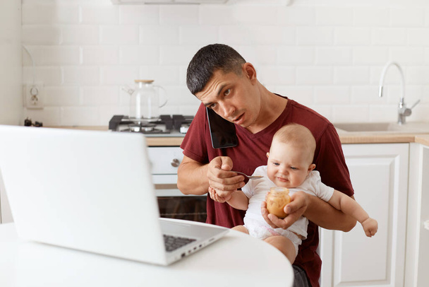 Indoor shot of busy brunette male wearing maroon casual style t shirt sitting at table in kitchen feeding his infant daughter with fruit puree, talking via cell phone looking at laptop display. - Φωτογραφία, εικόνα