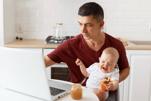 Concentrated attractive brunette man freelancer wearing casual style maroon t shirt, working on laptop and looking after his little baby daughter, sitting in white kitchen. - Valokuva, kuva