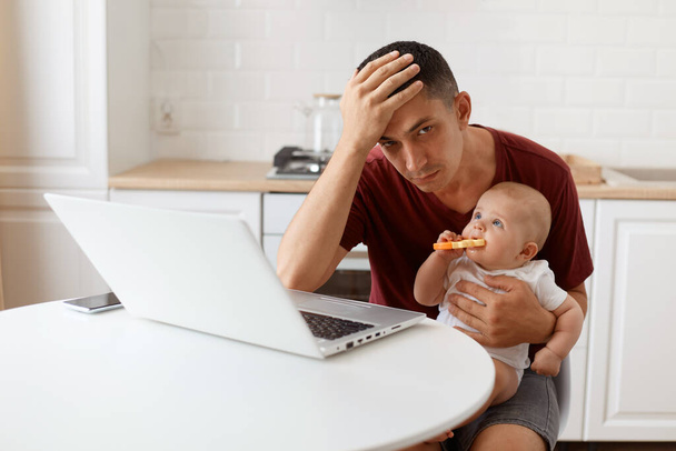 Exhausted attractive brunette man freelancer wearing casual style maroon t shirt, working and looking after his little baby daughter, keeping hand on his forehead. - Foto, immagini