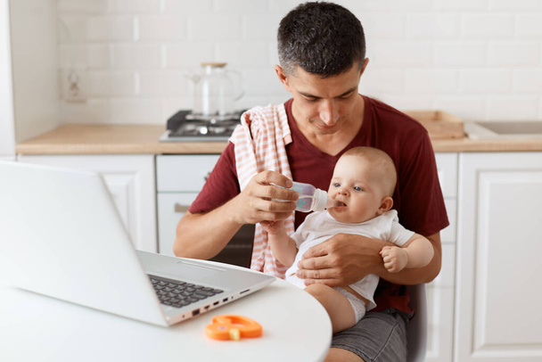 Indoor shot of dark haired handsome man wearing casual t shirt with towel on his shoulder, sitting at table with laptop, holding baby girl in hands and giving water from bottle to his daughter. - Valokuva, kuva