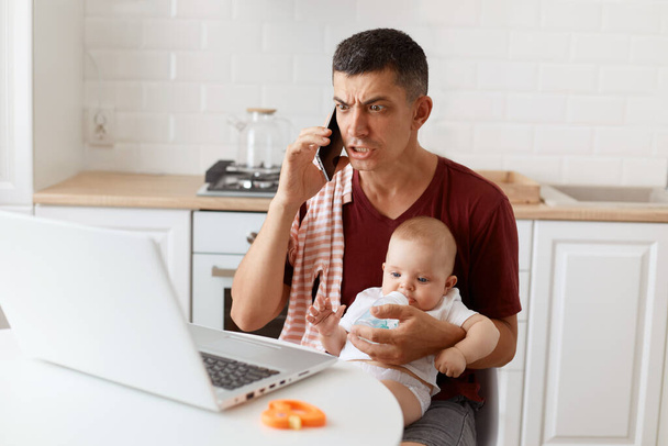 Scared serious man wearing burgundy casual t shirt with towel on his shoulder, looking after baby and working online from home, talking phone with partner, having troubles on work. - Fotoğraf, Görsel