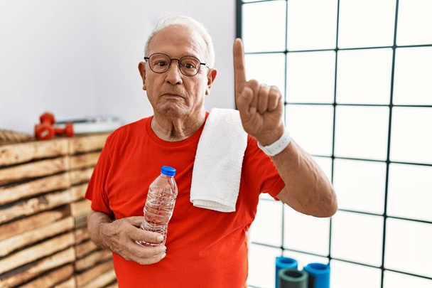 Hombre mayor con ropa deportiva y toalla en el gimnasio apuntando con el dedo hacia arriba y expresión enojada, sin mostrar ningún gesto  - Foto, Imagen