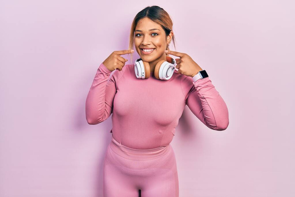 Beautiful hispanic woman wearing gym clothes and using headphones smiling cheerful showing and pointing with fingers teeth and mouth. dental health concept.  - Foto, Bild