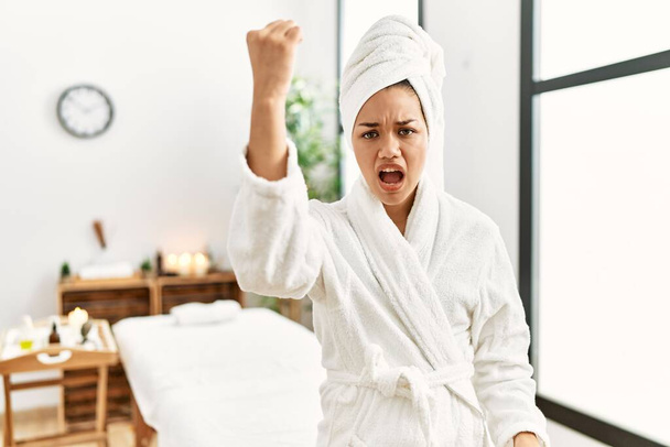 Young brunette woman wearing towel and bathrobe standing at beauty center angry and mad raising fist frustrated and furious while shouting with anger. rage and aggressive concept.  - Foto, Imagem