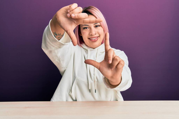Spaanse vrouw met roze haar in casual sweatshirt zittend op de tafel glimlachend het maken van frame met handen en vingers met gelukkig gezicht. creativiteit en fotografie.  - Foto, afbeelding