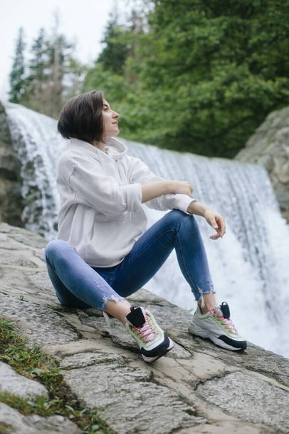 Mujer viajera sentada en una roca frente a una cascada en lo alto de las montañas, espacio para el texto, momento épico atmosférico - Foto, imagen