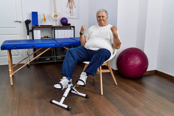 Старший кавказький чоловік у фізіотерапевтичній клініці використовує педальні вправи, які святкують здивовані та вражені успіхом з піднятими руками та відкритими очима. концепція переможця
.  - Фото, зображення
