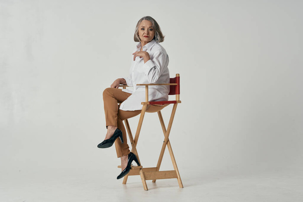 an elderly woman in a white shirt sits on a chair posing - 写真・画像