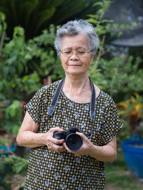 Femme âgée tenant un appareil photo numérique debout dans un jardin. Femme asiatique âgée cheveux blancs courts, portant des lunettes, heureux lors de l'utilisation d'un appareil photo. Concept de personnes âgées et photographie - Photo, image