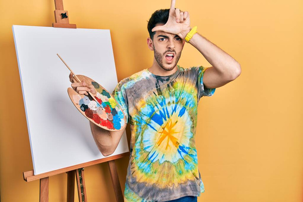 Junger hispanischer Mann, der mit einer Palette am Staffelei-Stand des Malers zeichnet und sich über Menschen mit Fingern auf der Stirn lustig macht, macht sich über Verlierer lustig und beleidigt.  - Foto, Bild