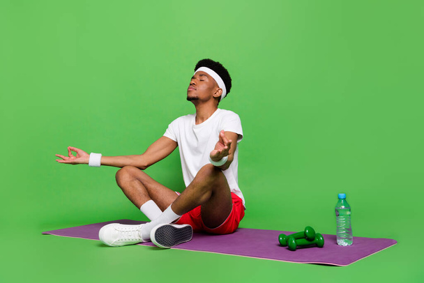 Full length photo of cute dreamy dark skin man wear white t-shirt practicing yoga sitting mat isolated green color background - Foto, afbeelding