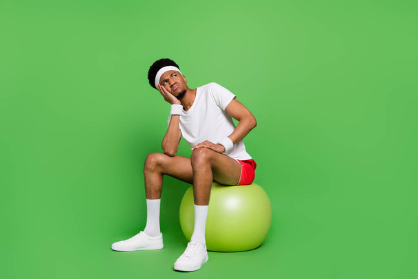 Full length body size view of trendy minded skeptic guy sportsman sitting on ball thinking isolated over bright green color background - Foto, afbeelding
