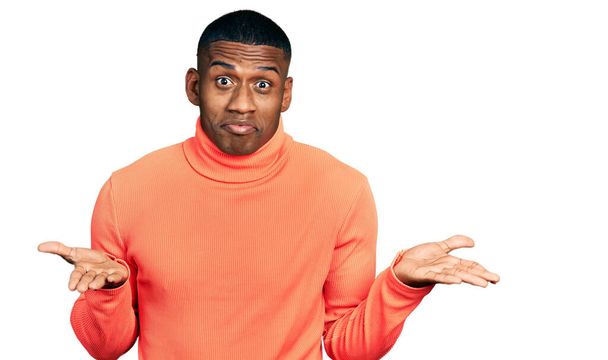 Молодой черный мужчина в оранжевой водолазке свитер невежественное и запутанное выражение лица с поднятыми руками и руками. Концепция сомнений.  - Фото, изображение