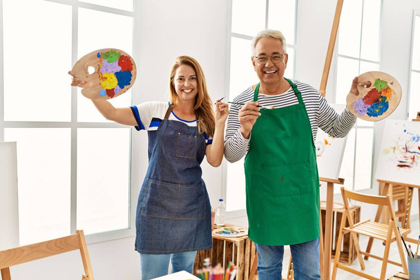 Dwóch artystów w średnim wieku uśmiechających się szczęśliwie trzymając pędzle i paletę w pracowni artystycznej. - Zdjęcie, obraz