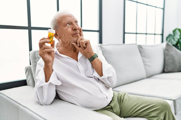 Senior man holding pills with hand on chin thinking about question, pensive expression. smiling and thoughtful face. doubt concept.  - Valokuva, kuva