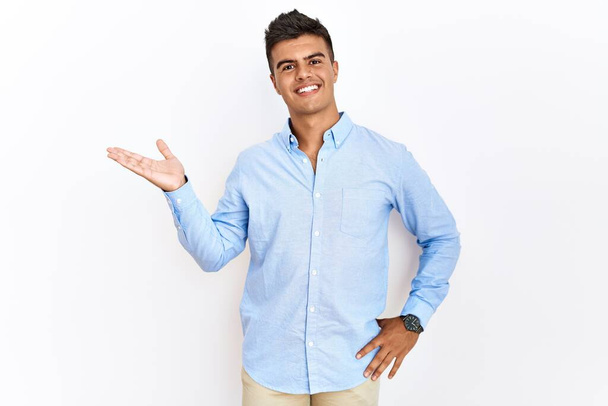 Jeune homme hispanique portant chemise d'affaires debout sur fond isolé souriant gai présentation et pointant avec la paume de la main regardant la caméra.  - Photo, image