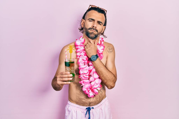 Young hispanic man wearing swimsuit and hawaiian lei drinking tropical cocktail touching painful neck, sore throat for flu, clod and infection  - Photo, Image
